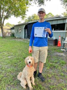 Man holding a Canine Good Citizen certificate and ribbon with his labradoodle standing next to him.